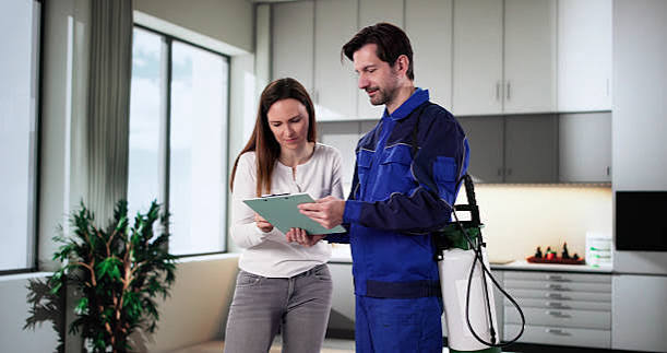 Pest-Control Man in a pest control uniform looking at a clipboard and talking to a woman in a kitchen.